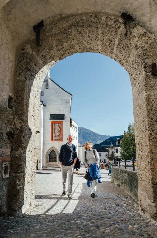 Coppia sorridente che cammina sotto un arco di pietra in una città storica europea con montagne sullo sfondo